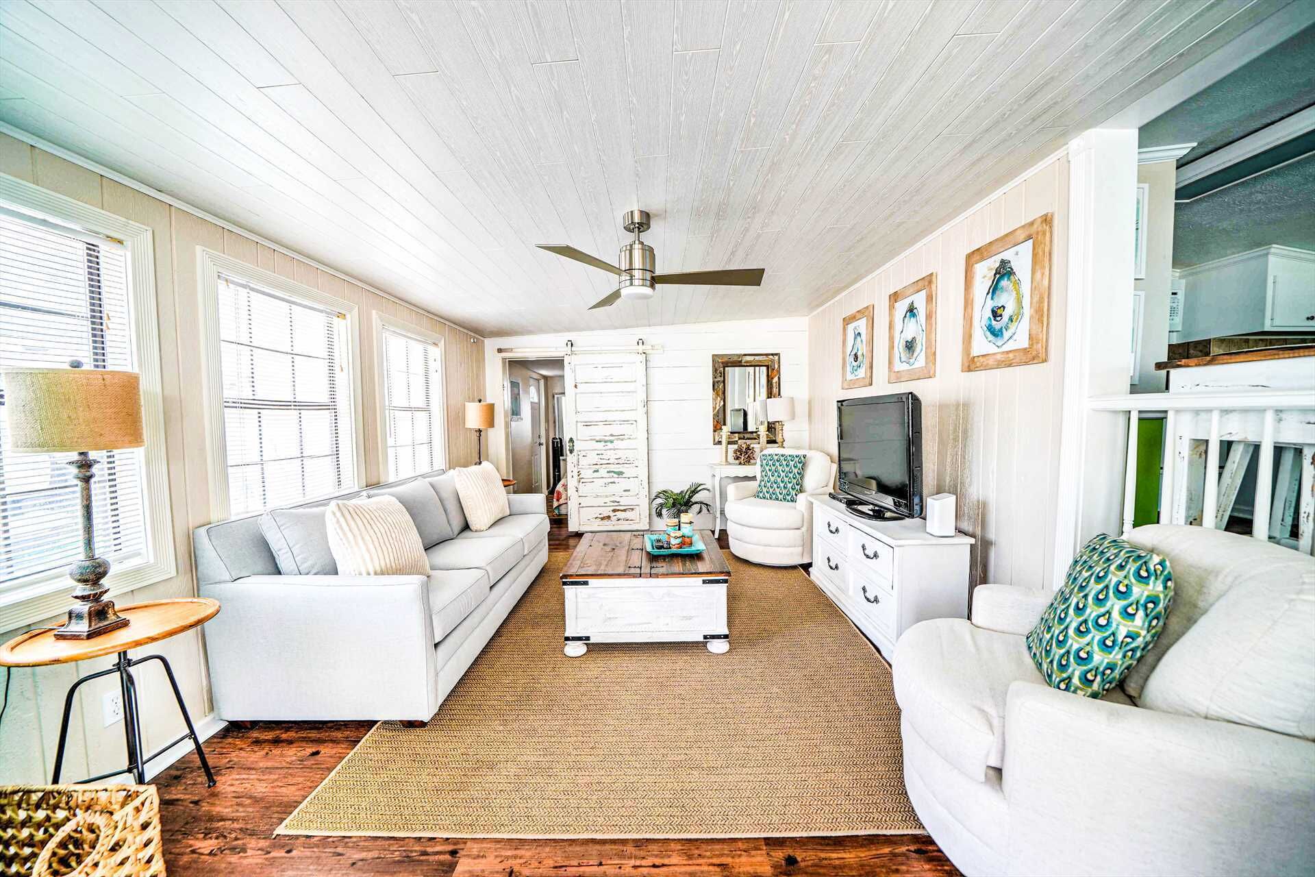 A spacious living room with a white sofa, a coffee table, and a TV. There are framed pictures on the wall and a ceiling fan.