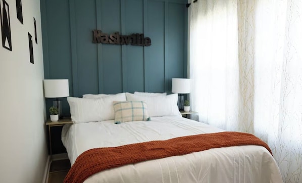 A bedroom with a bed, two bedside tables, a nightstand, a wall-mounted lamp, and a window with curtains.