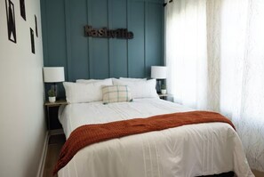 A bedroom with a bed, two bedside tables, a nightstand, a wall-mounted sign, and a window with curtains.