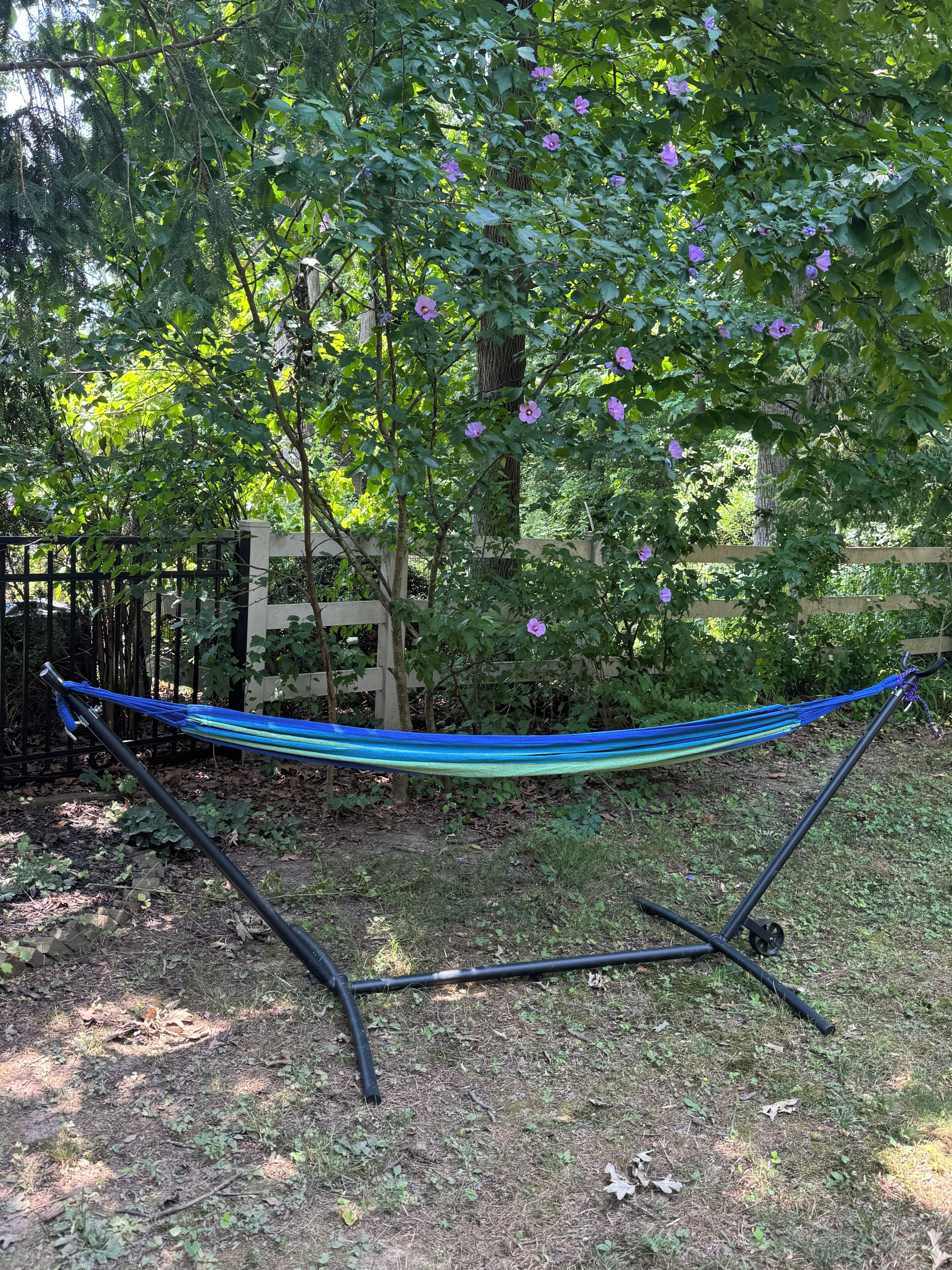 Een hangmat tussen twee palen in een tuin met bomen en een houten schutting.