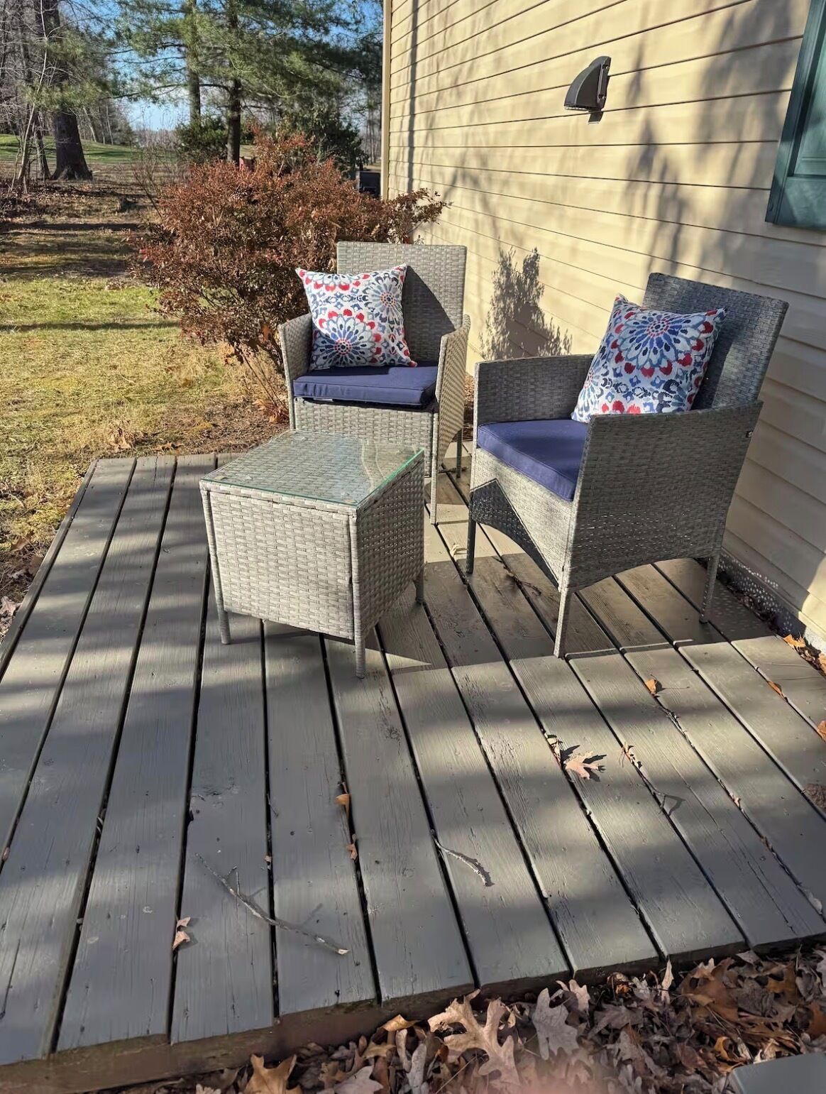 A covered deck with wicker furniture and cushions, a glass table, and a house in the background.
