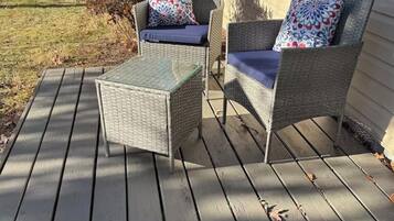 A covered deck with wicker furniture and cushions, a glass table, and a house in the background.