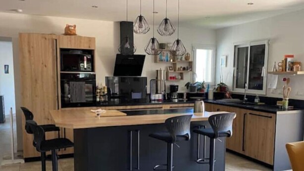 Una cocina moderna con una isla central, taburetes de bar, iluminación colgante y un ventanal grande.
