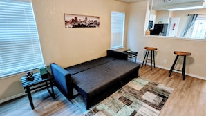 A compact living space with a sofa bed, a small table, two bar stools, and a cityscape painting on the wall. - Cozy 2-bedroom home, North East San Antonio (San Antonio)