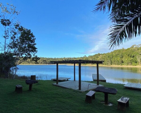 A gazebo with benches by a lake, surrounded by grass and trees. - Vila Tucunaré Chalés Premium - Camaçari (Camaçari)