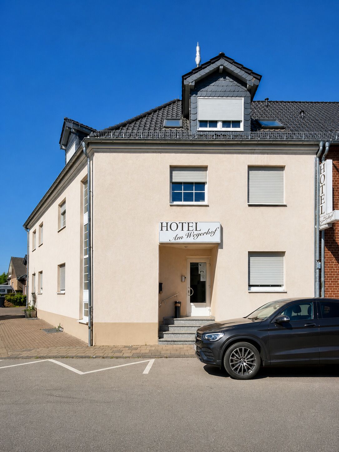 Een hotel met een bord waarop 'HOTEL Am Weyerhof' staat, een auto ervoor geparkeerd, en een heldere blauwe lucht.