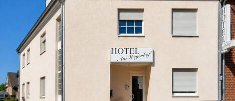 A hotel with a sign reading 'HOTEL Am Weyerhof,' a car parked in front, and a clear blue sky.