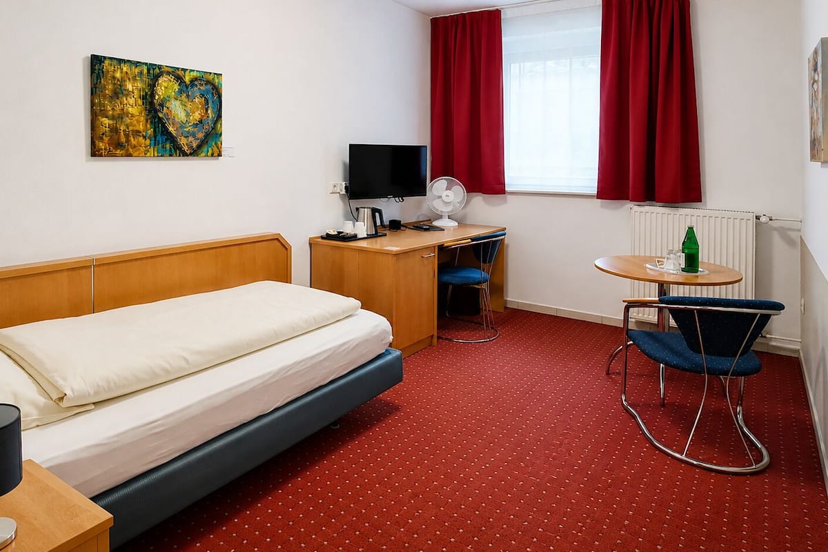 Una camera d'albergo con un letto, una scrivania con un computer, una sedia, una TV e una finestra con tende rosse.