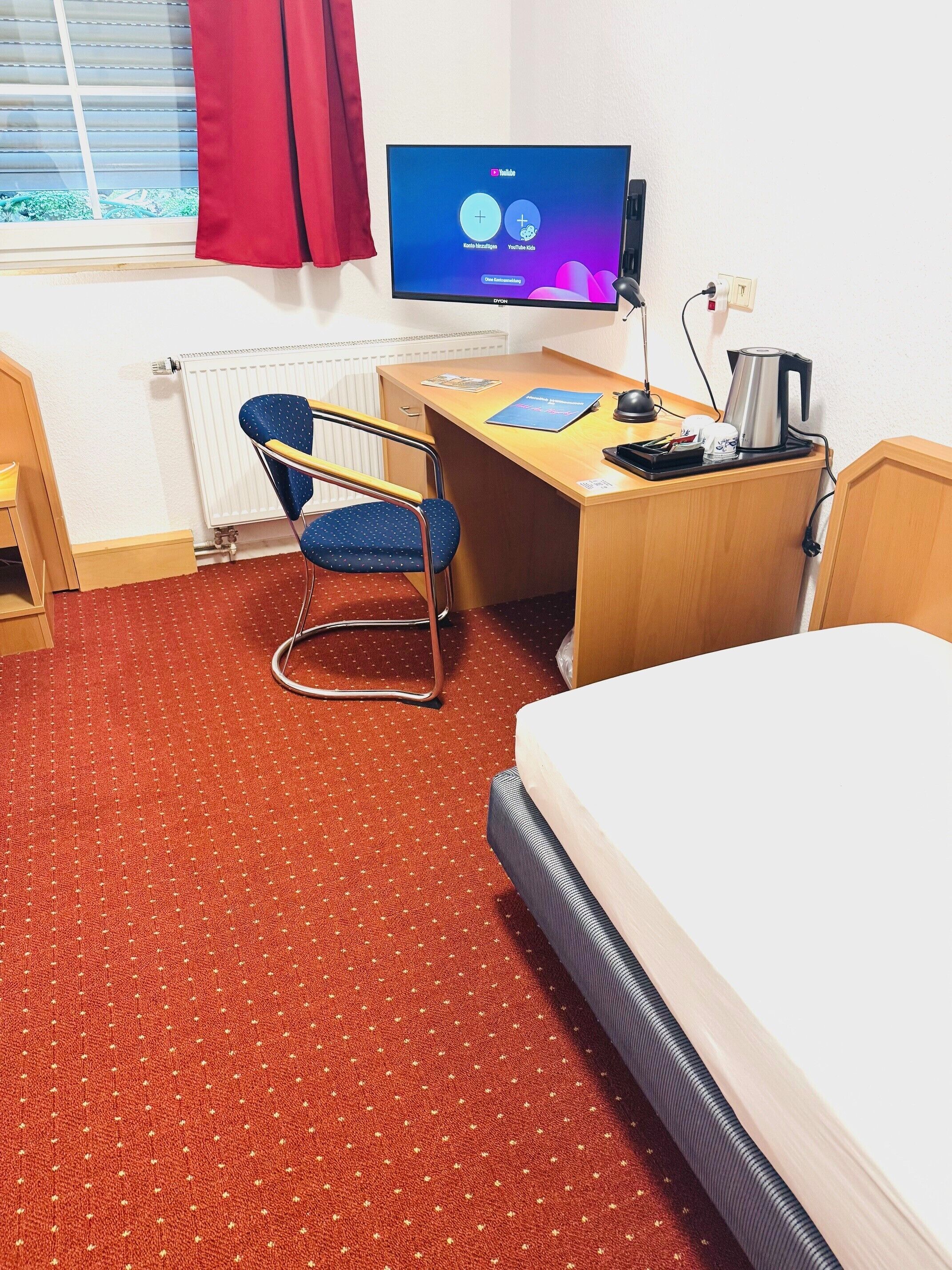A hotel room with a bed, a desk, a chair, a TV, and a radiator.