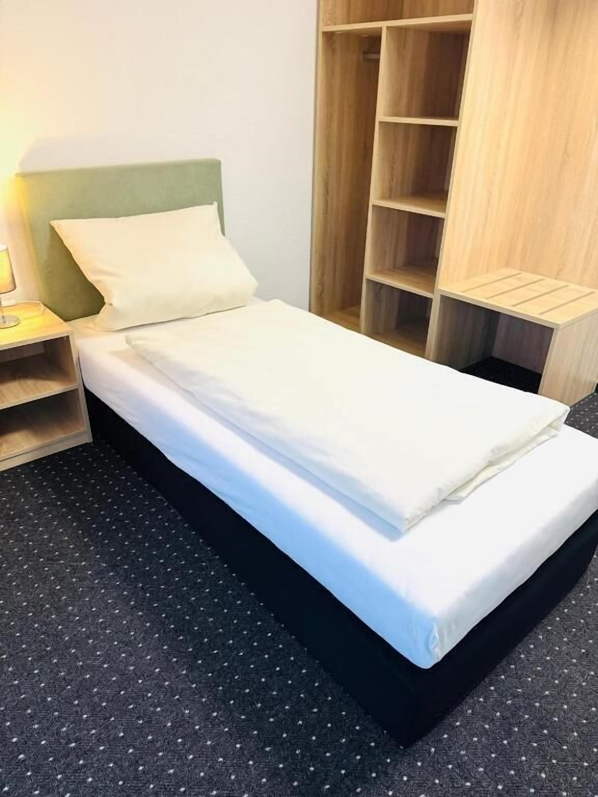 A single bed with a green headboard, a bedside table, and a wooden wardrobe with multiple shelves.
