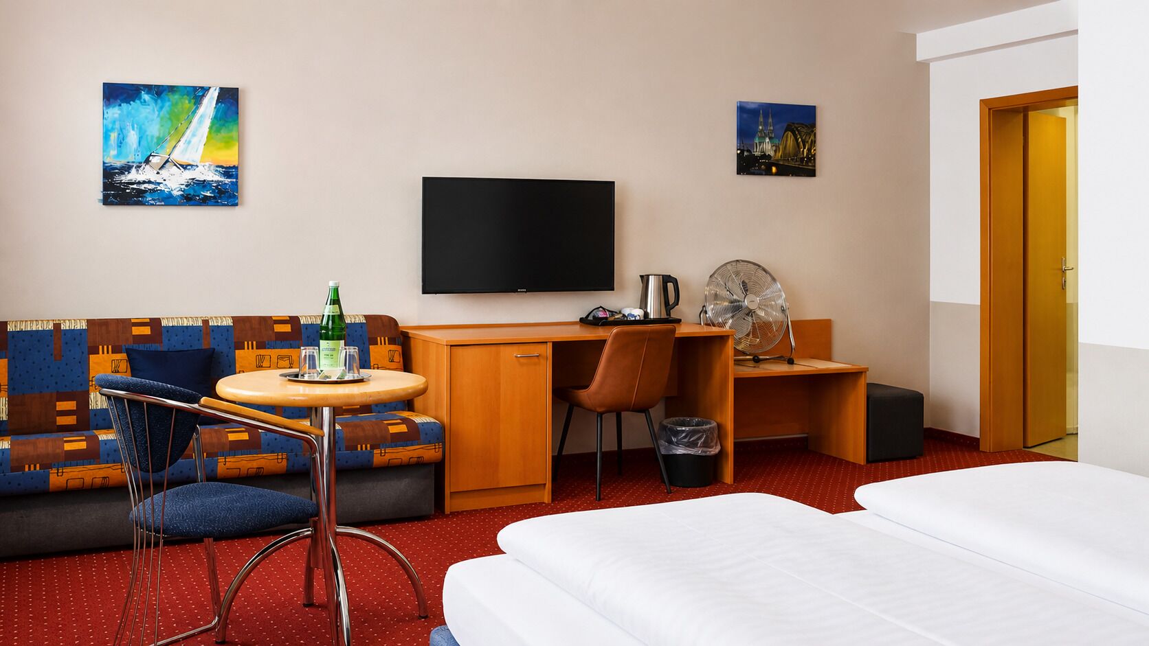 A hotel room with a bed, a sofa, a desk, a TV, and artwork on the walls.