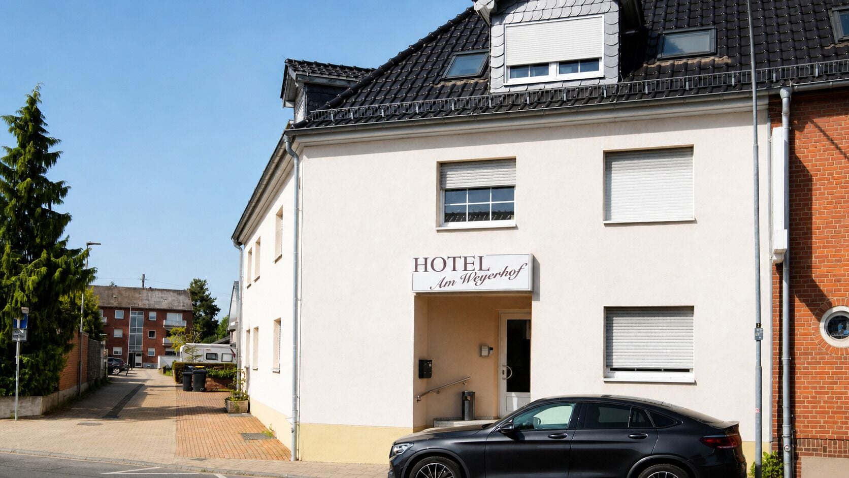 Hotellin edessä on kyltti, jossa lukee ”HOTEL Am Weyerhof”, asfaltoitu ajotie ja parkissa oleva auto.