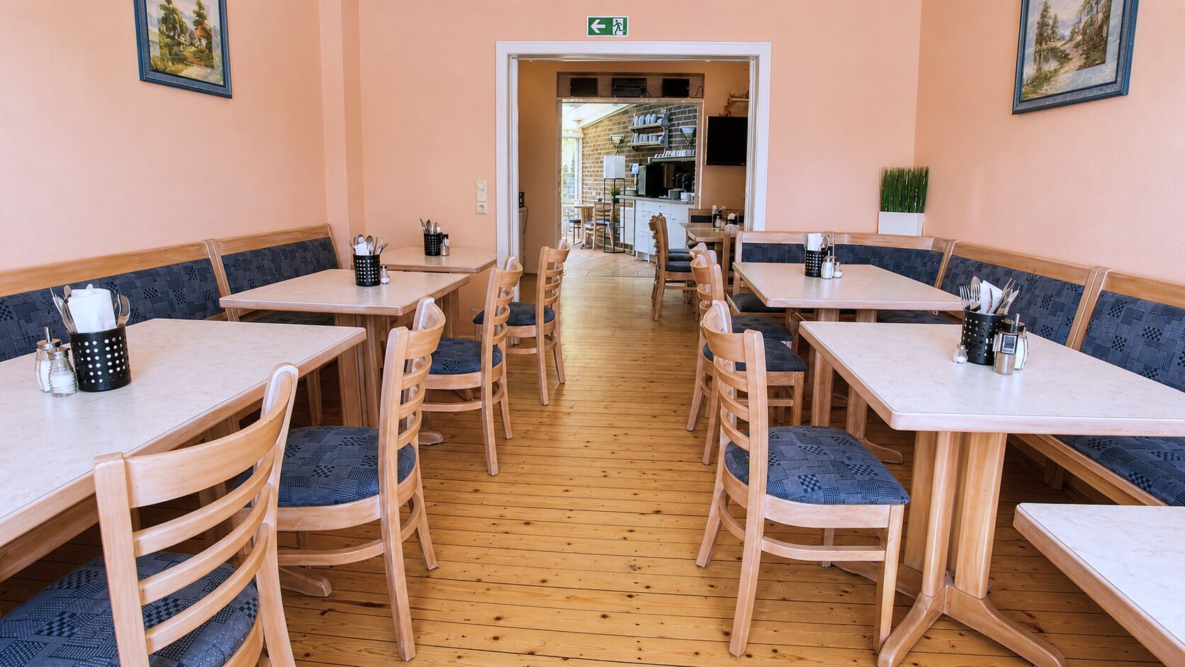 A restaurant with wooden tables and chairs, a tiled floor, and a view of the kitchen in the background.
