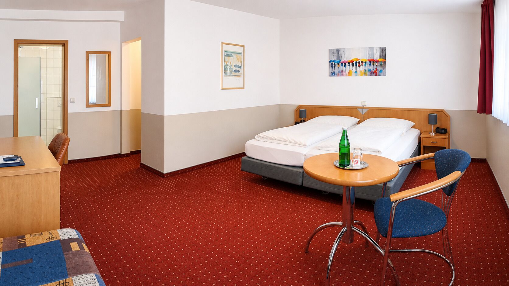 A hotel room with a bed, a desk, a chair, a small table, and artwork on the walls.