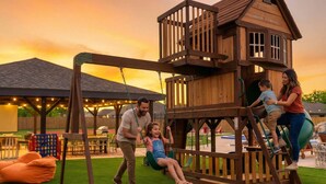 Eine hölzerne Spielkonstruktion mit Schaukel und Rutsche, umgeben von Kunstgrün und Gartenmöbeln.