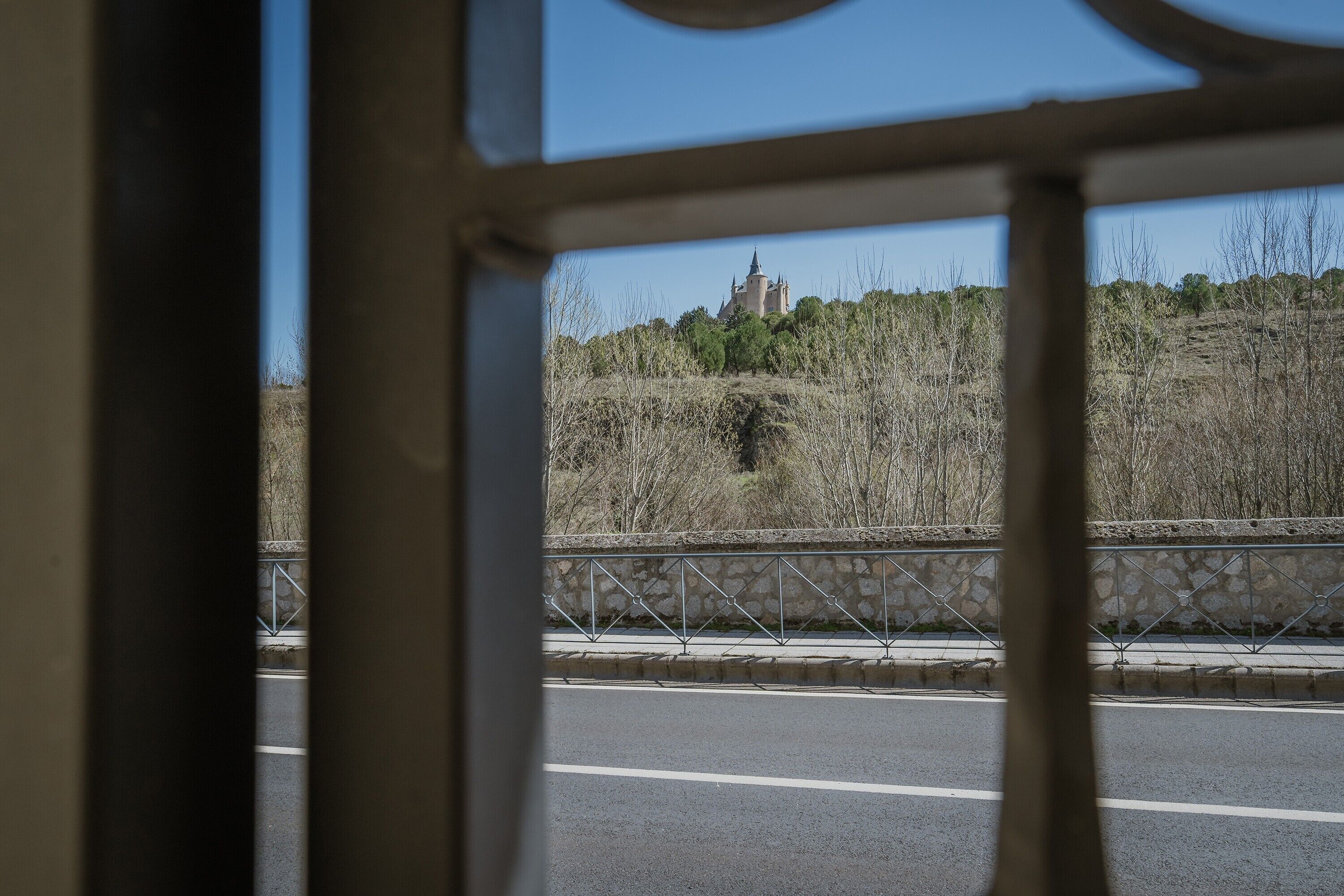 Vista a través de una ventana de un edificio con pared de piedra y barandilla de metal, una carretera en primer plano y una colina distante con una estructura.