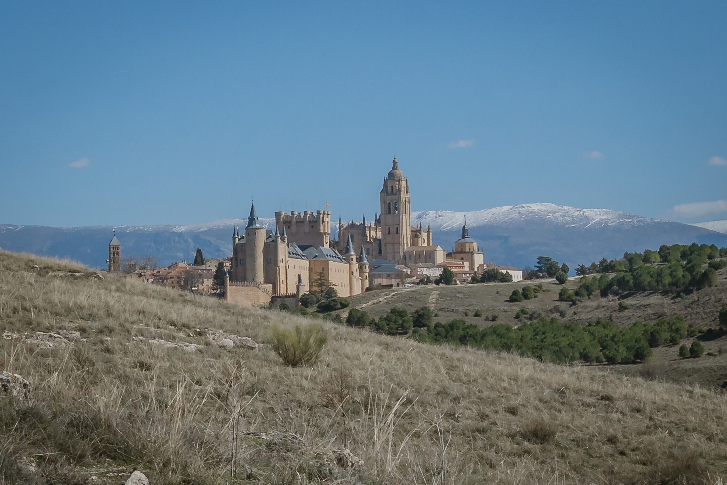 A historic castle with multiple towers and a prominent bell tower, surrounded by a hilly landscape with sparse vegetation.