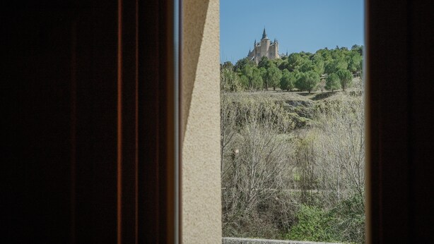 En vy genom ett fönster, med ett slott i fjärran, omgivet av träd och ett kuperat landskap.
