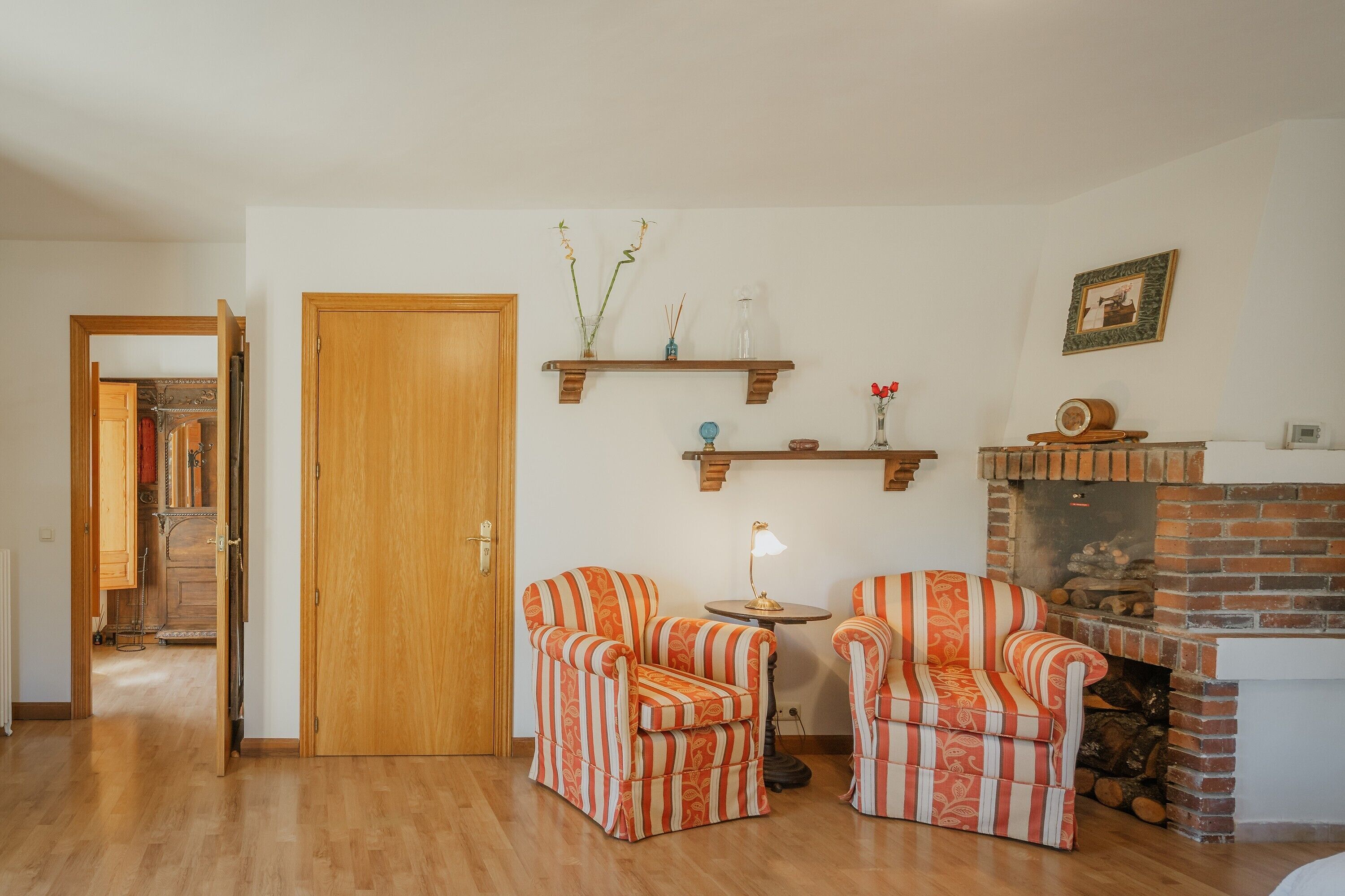 Een woonkamer met twee gestreepte fauteuils, een kleine tafel met een lamp, een open haard en een houten deur.
