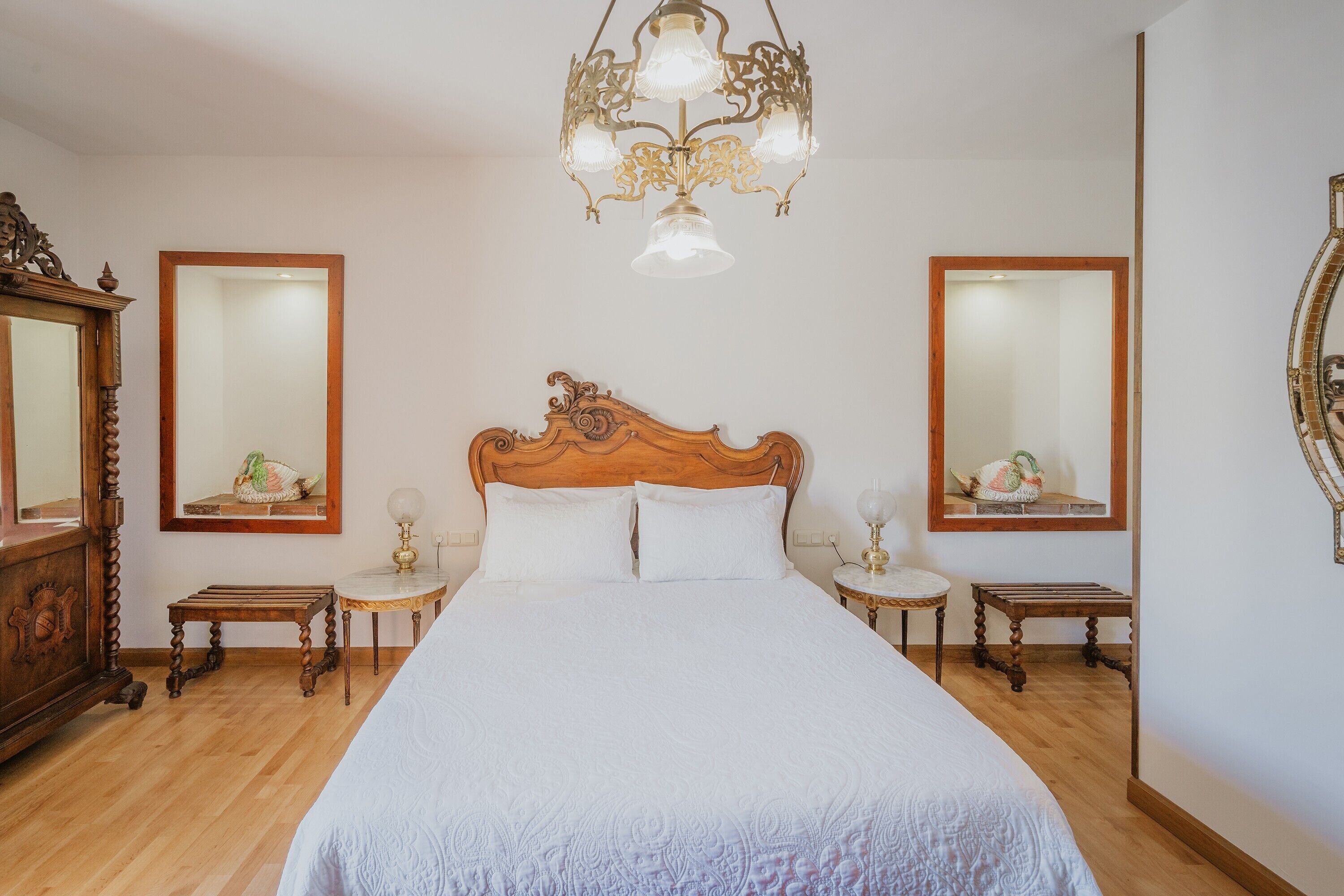 Ein Schlafzimmer mit einem hölzernen Bett, zwei Nachttischen, einer Kristallleuchtre und zwei Wandspiegeln.