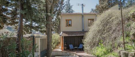 A house with a patio, two chairs, a small table, and a garden with trees and shrubs.