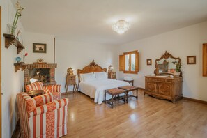 A bedroom with a bed, a wooden dresser, a chair, a bench, a fireplace, and a chandelier. - Rural House 'Casa Agapito Marazuela' with Mountain Views, Private Garden, and Wi-Fi (Segovia)