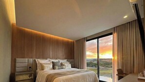 A modern bedroom with a large bed, a nightstand, and a view of the sunset through the window. - Moto GP Goiânia - Casa Premium - Piscina (Senador Canedo)