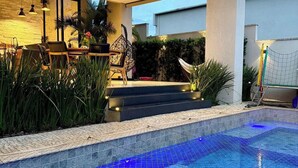 A modern outdoor pool area with a tiled floor, surrounded by a low wall and steps leading up to a covered seating area with chairs and a table. - Moto GP Goiânia - Casa Premium - Piscina (Senador Canedo)