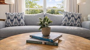 Een woonkamer met een grijze bank, twee gestreepte kussens, een houten bijzettafel met boeken en een potplant.