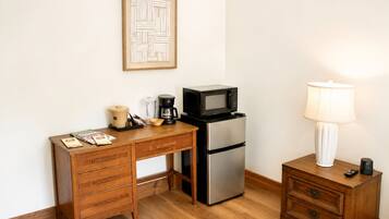 Una kitchenette compacta con escritorio de madera, refrigerador de acero inoxidable, microondas, cafetera y mesita de noche con lámpara.
