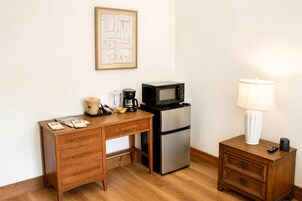 Una kitchenette compacta con escritorio de madera, refrigerador de acero inoxidable, microondas, cafetera y mesita de noche con lámpara.