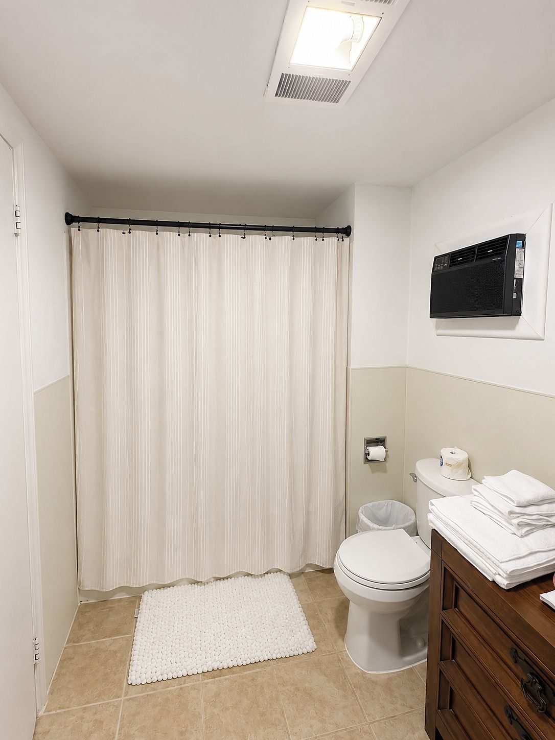 A bathroom with a toilet, a white shower curtain, a white rug, and a wall-mounted air conditioner.