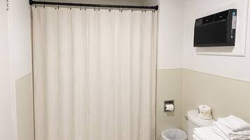 A bathroom with a toilet, a white shower curtain, a white rug, and a wall-mounted air conditioner.