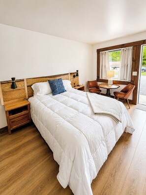 A hotel room with a large bed, a wooden headboard, a nightstand, a table with chairs, and a window with curtains.