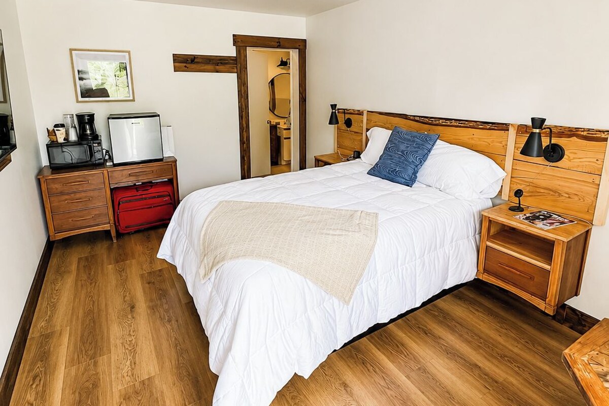 A bedroom with a wooden floor, a bed with white bedding, a nightstand with a lamp, a dresser, and a red suitcase.