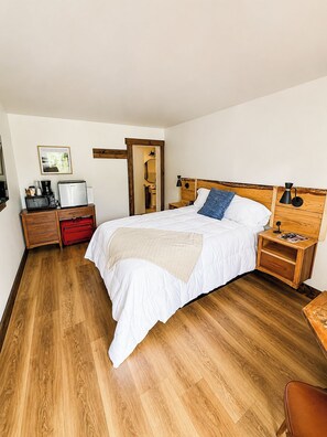 A bedroom with a wooden floor, a bed with white bedding, a nightstand with a lamp, a dresser, and a red suitcase. - Blue Ridge Mountain Inn (Meadows Of Dan)