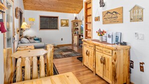 A rustic cabin interior with a wooden dining table, a kitchen area with wooden cabinets, a fireplace, and a cozy seating area with cushions.