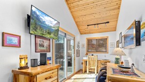 A cozy room with a wooden ceiling, a large TV mounted on the wall, a desk with a chair, and a window with blinds.
