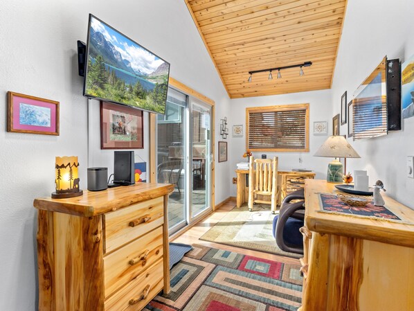 A cozy room with a wooden ceiling, a large TV mounted on the wall, a desk with a chair, and a window with blinds.