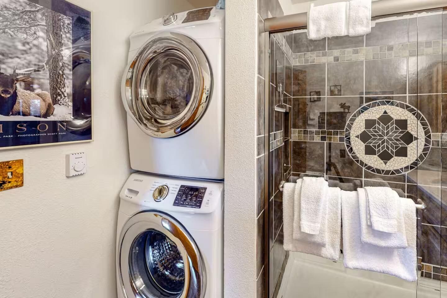 Un espace buanderie avec des machines à laver superposées et une douche en verre agrémentée de motifs décoratifs en carreaux.