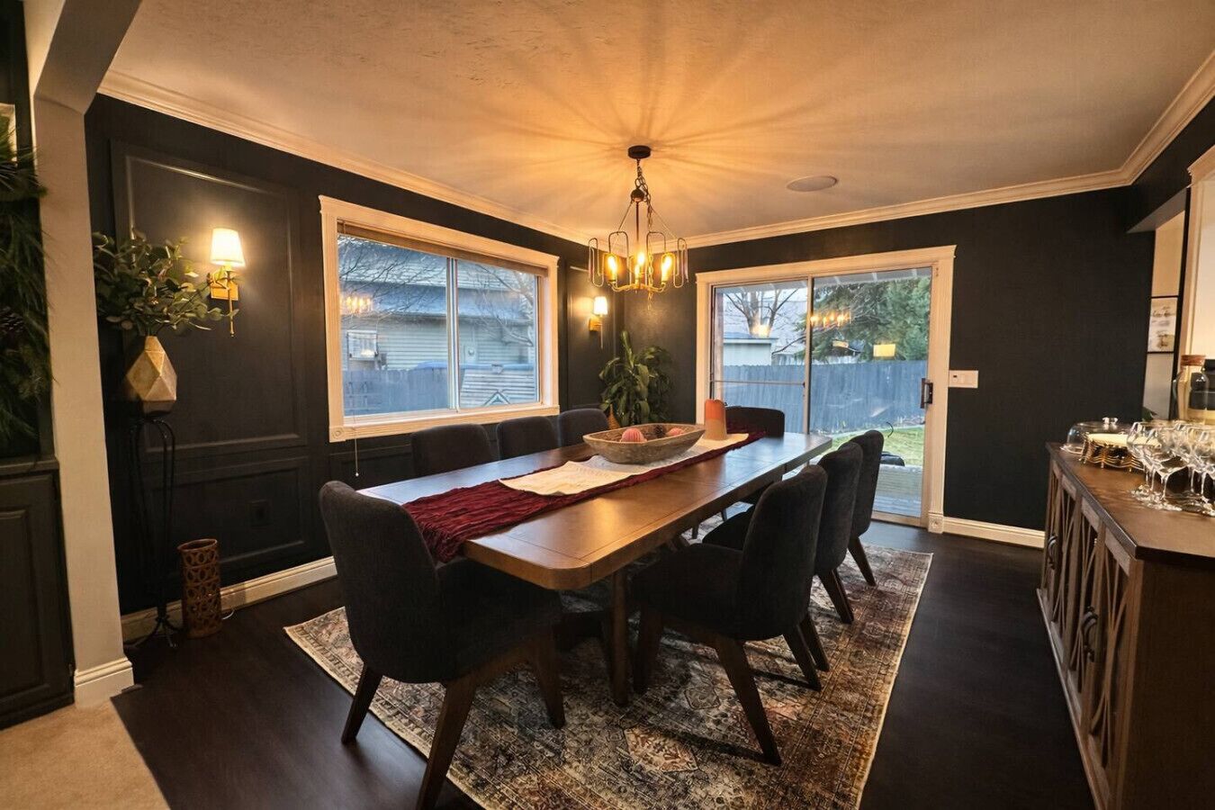 Un coin repas avec une table en bois, des chaises et un lustre. Il y a des fenêtres et une vue sur l’extérieur.