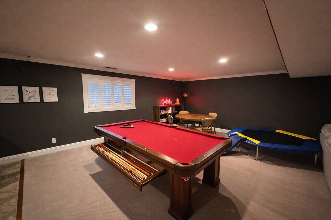 Uma mesa de bilhar com superfície em feltro vermelho, uma pequena mesa com cadeiras e um trampolim em um ambiente com paredes escuras e iluminação embutida no teto.