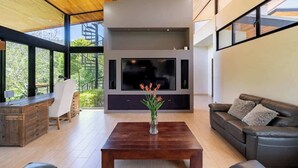 A modern living room with a large flat-screen TV, wooden ceiling, and a dining area with a wooden table and chairs.