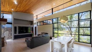 A modern living room with a large TV, a dining area with a white table and chairs, and a large window offering a view of trees.