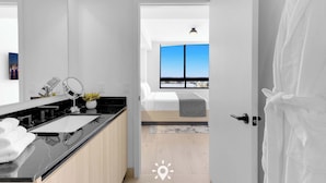 Un baño moderno con encimera negra, un lavabo y un espejo. Hay un perchero con toallas dobladas. El baño da acceso a un dormitorio con cama, ventana con vistas a la ciudad y balcón.