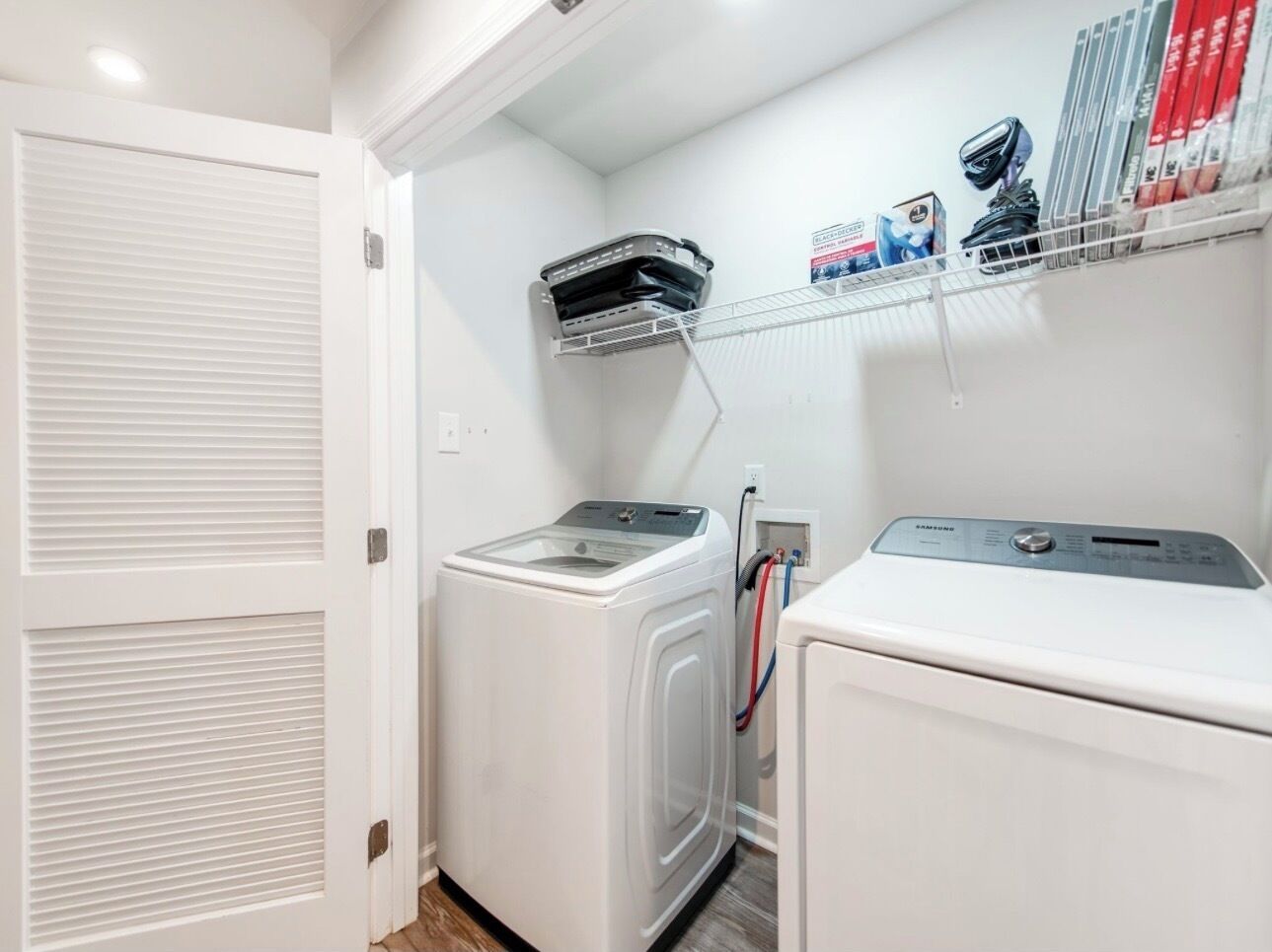 A laundry room with two white washing machines, a shelf with books and a camera, and a door with louvered panels.