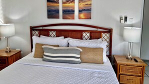 Una cama bien hecha con ropa blanca, cabecera de madera y tres cuadros de atardecer colgados en la pared.