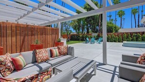 Een terras buiten met een witte pergola, een grijze bank met decoratieve kussens en een zwembad op de achtergrond.