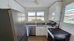 Una cocina moderna con electrodomésticos de acero inoxidable, armarios blancos, piso de madera y una ventana con persianas.