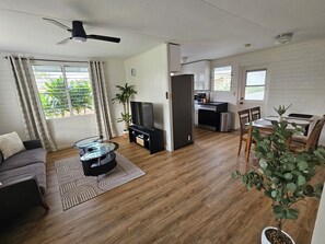 Ein modernes Wohnzimmer mit Sofa, Fernseher und Couchtisch, eine Küche mit Geräten sowie ein Essbereich mit Stühlen und Tisch.